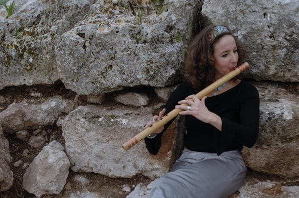 Tal Zimra Coleman playing a wooden flute against an ancient stone wall